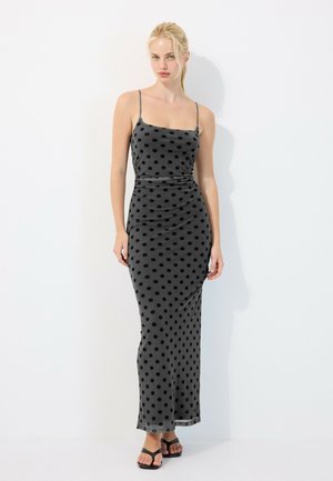 Femme blonde portant une longue robe grise à pois noirs et fines bretelles, debout sur un fond blanc.
