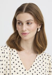 Femme aux cheveux bruns mi-longs portant une robe crème à pois noirs et des boucles d'oreilles argentées en forme de cercle, regardant légèrement vers sa gauche.
