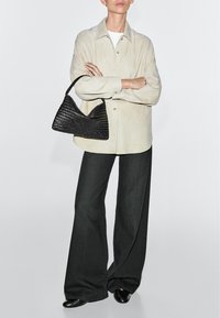 Person trägt ein beige Hemd mit Knöpfen, schwarze weit geschnittene Hosen, schwarze Schuhe und hält eine schwarze gewebte Handtasche, die Arme verschränkt.