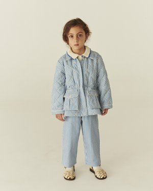 Ungt barn står, iført blå og hvide ternede, quiltede jakke og bukser med cremefarvet krave og vævede cremefarvede sko på ensfarvet baggrund.