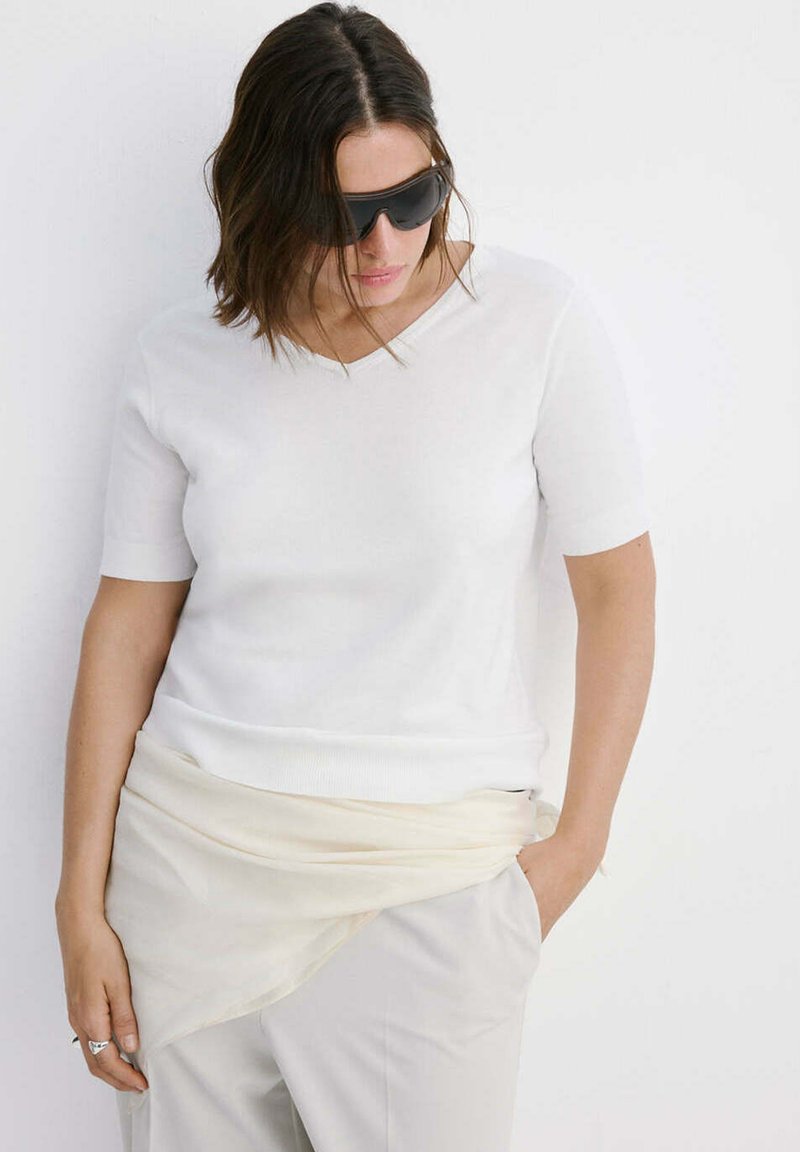 Femme portant des lunettes de soleil noires, un haut blanc à manches courtes, une jupe portefeuille beige clair et un pantalon clair, debout contre un mur blanc.