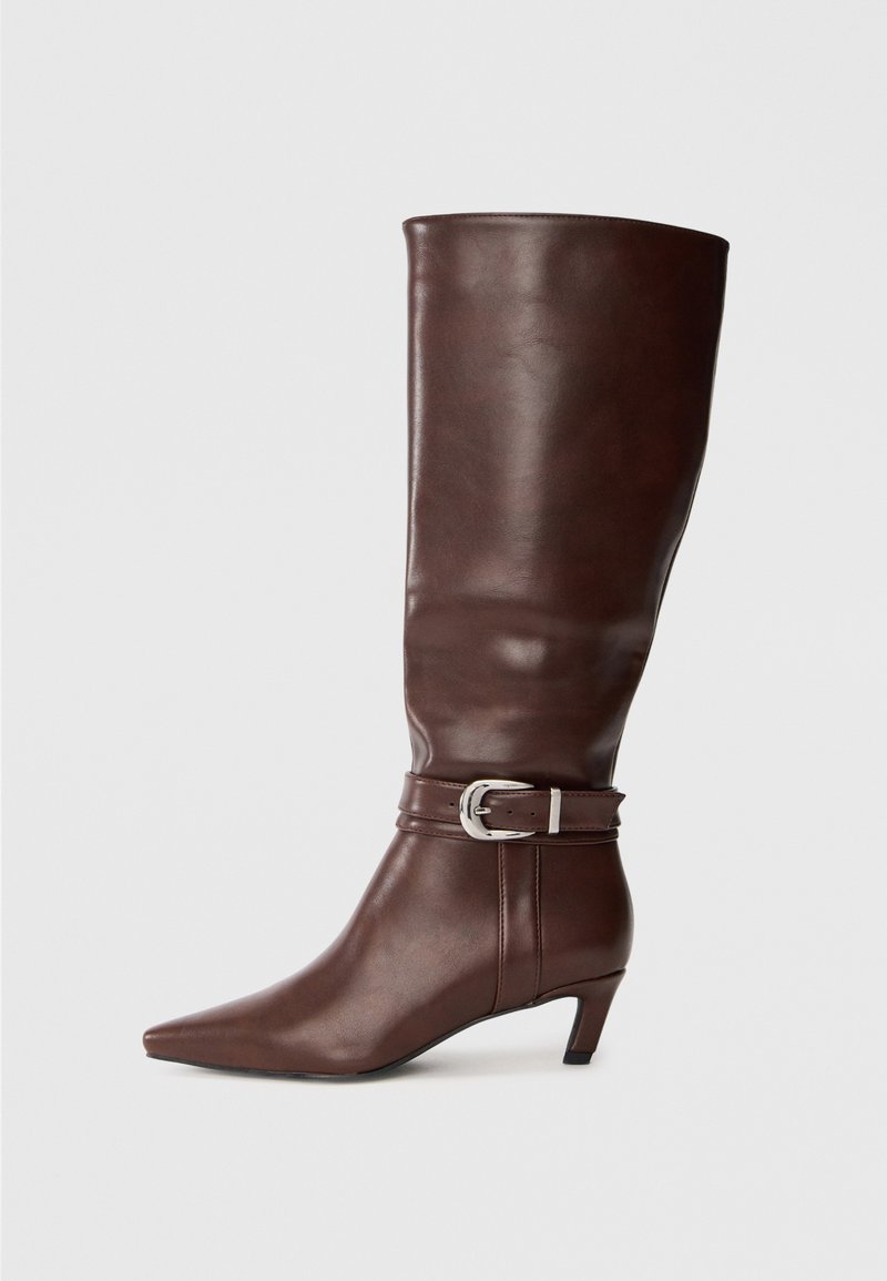 Bota de cuero marrón hasta la rodilla con punta afilada, tacón bajo y tira decorativa con hebilla alrededor del tobillo.