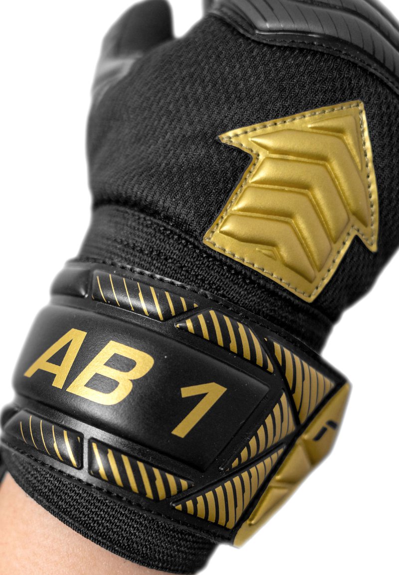 Black sports glove with gold accents, featuring an embossed arrow design and "AB 1" label. Texture is a mix of fabric and padded elements.