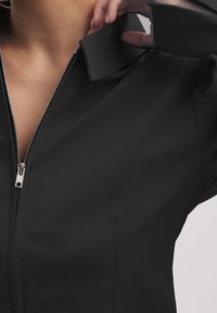 Nahaufnahme einer Person in einer schwarzen zip-up Jacke, die mit der Hand den Kragen nahe am Hals anpasst.