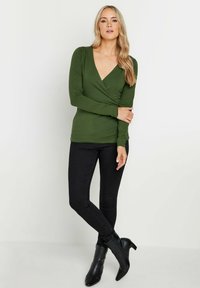 Top avvolgente verde oliva a maniche lunghe con scollo a V profondo, abbinato a jeans skinny neri e stivaletti, che presenta un design elegante e aderente.