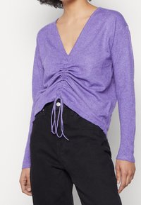 Fioletowy sweter z dekoltem w serek, z marszczeniami w talii i ozdobnymi sznureczkami, wykonany z miękkiej, teksturowanej tkaniny.