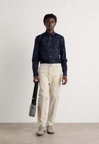 Camisa de manga larga azul marino, pantalones beiges, bolso cruzado gris y zapatillas grises; la modelo se encuentra de pie contra un fondo neutral.