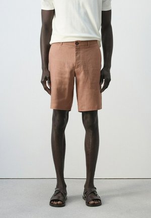 Modello maschile che indossa pantaloncini marrone chiaro fino al ginocchio, camicia bianca a maniche corte e sandali marrone scuro, in piedi su uno sfondo chiaro e semplice.