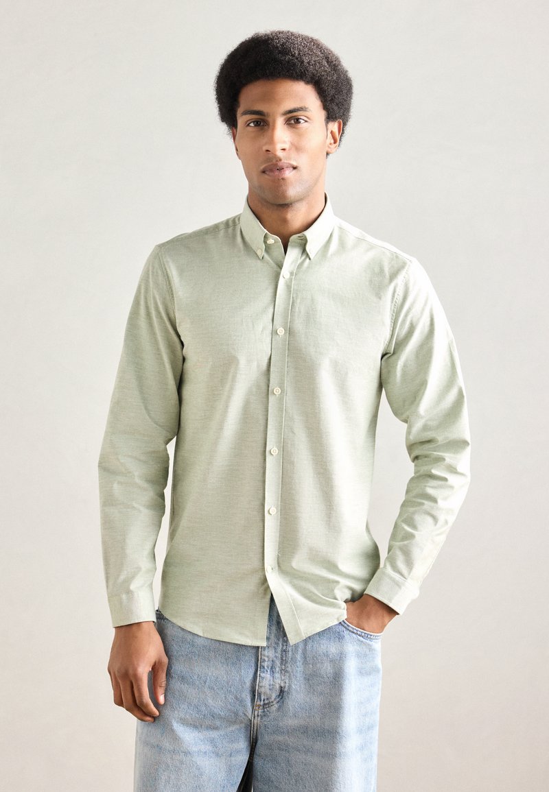 Ανοιχτοπράσινο πουκάμισο με μακριά μανίκια, κουμπιά και γιακά, υφασμένο με υφή και στρογγυλεμένη κάτω άκρη. Συνδυασμένο με ανοιχτό μπλε τζιν παντελόνι.