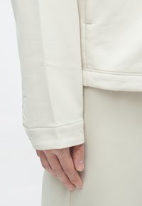 Manche de veste blanche ajustée en tissu texturé avec des détails de couture, associée à un vêtement de couleur claire unie. La main repose délicatement contre le tissu.