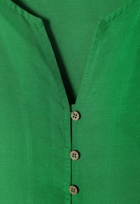 Top in tessuto verde con scollatura a V, caratterizzato da una lista di quattro bottoni rotondi in legno e una superficie liscia e testurizzata.