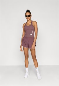 Αθλητικό τοπ Nike σε σκούρο μοβ με λευκό λογότυπο, σετ με αντίστοιχα σορτς με υφή και λευκή επένδυση. Μαύρα γυαλιά ηλίου.