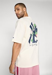 Cream T-shirt with a navy floral NY logo on the back. Short sleeves and a relaxed fit. Shirt paired with pink athletic shorts.