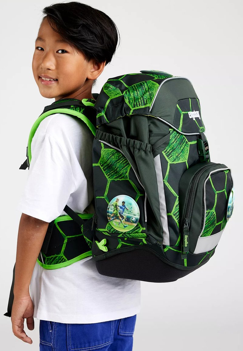 Garçon portant une chemise blanche et un pantalon bleu, portant un grand sac à dos noir et vert à motif hexagonal avec un écusson de joueur de football.