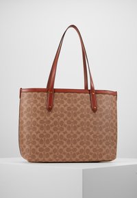 Sac fourre-tout marron avec un motif monogramme marron foncé, des bordures en cuir et des sangles doubles, exposé sur un piédestal blanc devant un fond gris.