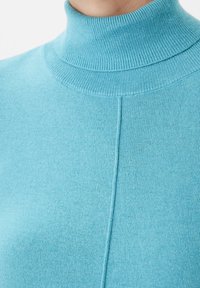 Primer plano de una persona que lleva un suéter de cuello alto de punto en color azul claro con un detalle de costura vertical en la parte delantera.