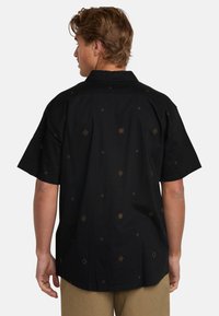 Camisa negra de manga corta hecha de algodón, con patrones geométricos dorados, cuello tipo polo y un corte relajado, vista desde atrás.