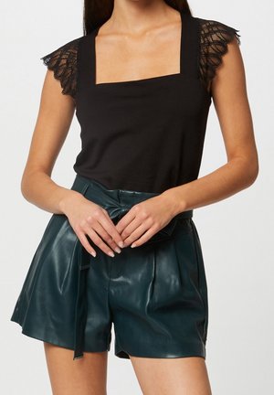 Vrouw draagt een zwarte mouwloze top met kanten schouders en donkerrode leren shorts met hoge taille, vastgebonden met een riem in de taille.