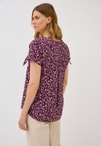 Blouse à manches courtes de couleur violet foncé avec un motif de feuilles. Présente des détails à nouer sur les manches et un ourlet arrondi. Tissu léger et doux.