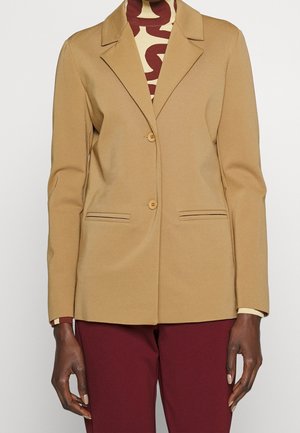 Camelowy blezer na dwóch guzikach noszony na wzorzystej bluzce z wysokim kołnierzem, w połączeniu z bordowymi spodniami, widoczny od szyi do bioder.