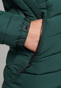 Chaqueta acolchada verde con bolsillo con cremallera; cuenta con textura acolchada, puño elástico y tirador de cremallera para cierre seguro.