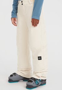 Beige hiihtohousut, joissa on rento malli, logopatchilla lahkeessa ja kestävästä kankaasta. Yhdistetty harmaisiin hiihtobootseihin, joissa on sinisiä yksityiskohtia.