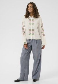 Weißes Langarmhemd mit mehrfarbigem Blumenstickerei an den Ärmeln, kombiniert mit grauen gestreiften weit geschnittenen Hosen und schwarzen spitzen Schuhen.