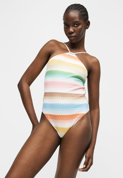 Γυναίκα που φοράει αμάνικο ολόσωμο μαγιό με πολύχρωμες οριζόντιες ζιγκ ζαγκ ρίγες, ποζάροντας μπροστά σε απλό φόντο.