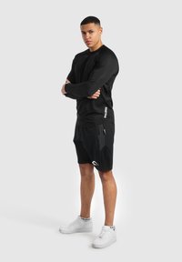 Schwarzes Langarmshirt und schwarze Shorts aus leichtem Material, mit dezentem Muster und Logoakzenten. Weiße Sportschuhe runden den Look ab.