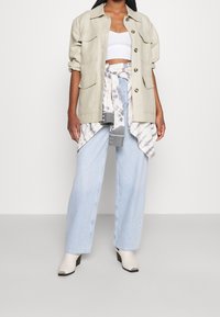 Veste utilitaire beige surdimensionnée avec plusieurs poches, portée sur un top court blanc et une chemise grise tie-dye nouée. Jean bleu clair à jambes larges et bottines blanches.