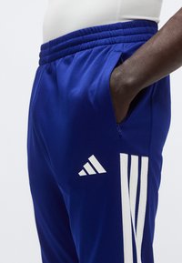 Μπλε αθλητικό παντελόνι με ελαστική μέση, πλαϊνή τσέπη με φερμουάρ και λευκό σχέδιο με τρεις ρίγες στο πόδι, που φέρει το λογότυπο της Adidas.