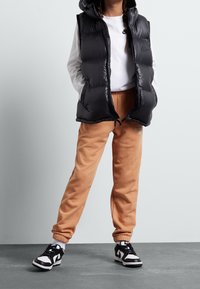 Chaleco puffer negro con capucha, camisa gris de manga larga, pantalones de chándal beige y zapatillas negras con detalles blancos. Presenta los logotipos de Nike y New Balance.