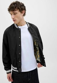 Veste bomber noire avec col côtelé rayé blanc, boutons-pression en argent et doublure à motifs floraux ; portée par-dessus un t-shirt blanc.