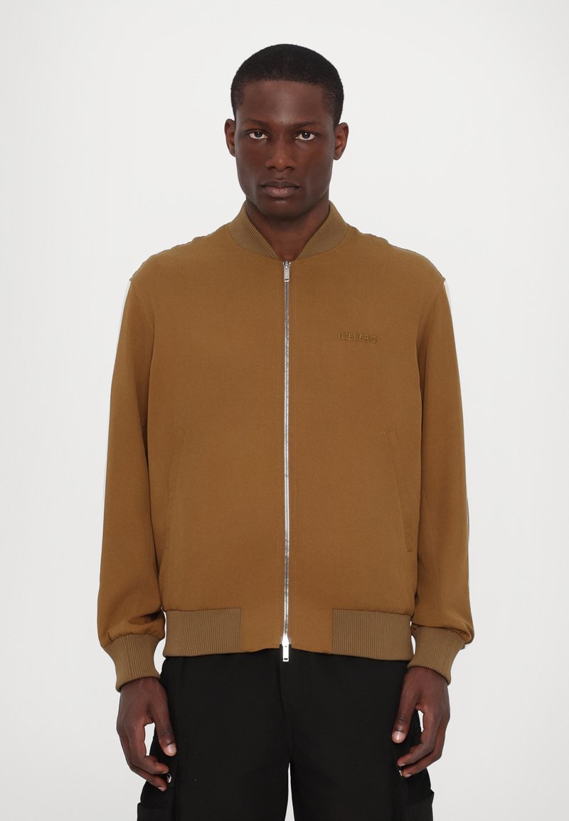 Veste bombardier marron avec fermeture éclair, bords côtelés aux poignets et à l'ourlet, dotée de poches latérales et d'un logo de marque discret sur la poitrine.