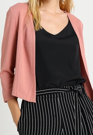 Femme portant un blazer rose, un top noir à col en V et un pantalon noir à rayures verticales blanches noué à la taille.