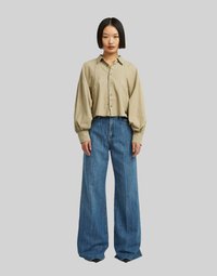 Maglietta beige cropped con bottone frontale e maniche a sbuffo abbinata a jeans in denim blu a gamba larga. Il tessuto appare morbido e testurizzato.