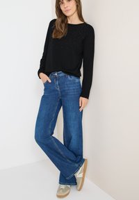 Kobieta opiera się o ścianę, ubrana w czarną bluzkę z długim rękawem ozdobioną małymi ćwiekami, niebieskie szerokie dżinsy i beżowe trampki.