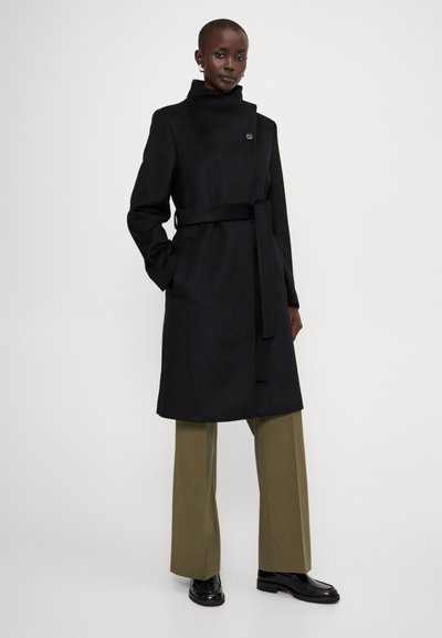 Un long trench noir avec un col haut et une ceinture, comportant une fermeture à un bouton et des poches latérales. Associé à un pantalon olive et des chaussures noires.