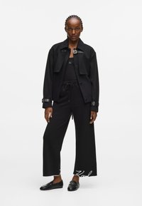 Melnā pārmērīga žakete ar sprādzi, uzvilkta virs pieguļošas melnas blūzes un melniem platām biksēm ar baltu tekstu. Izskatu papildina melnas mokasīnas.