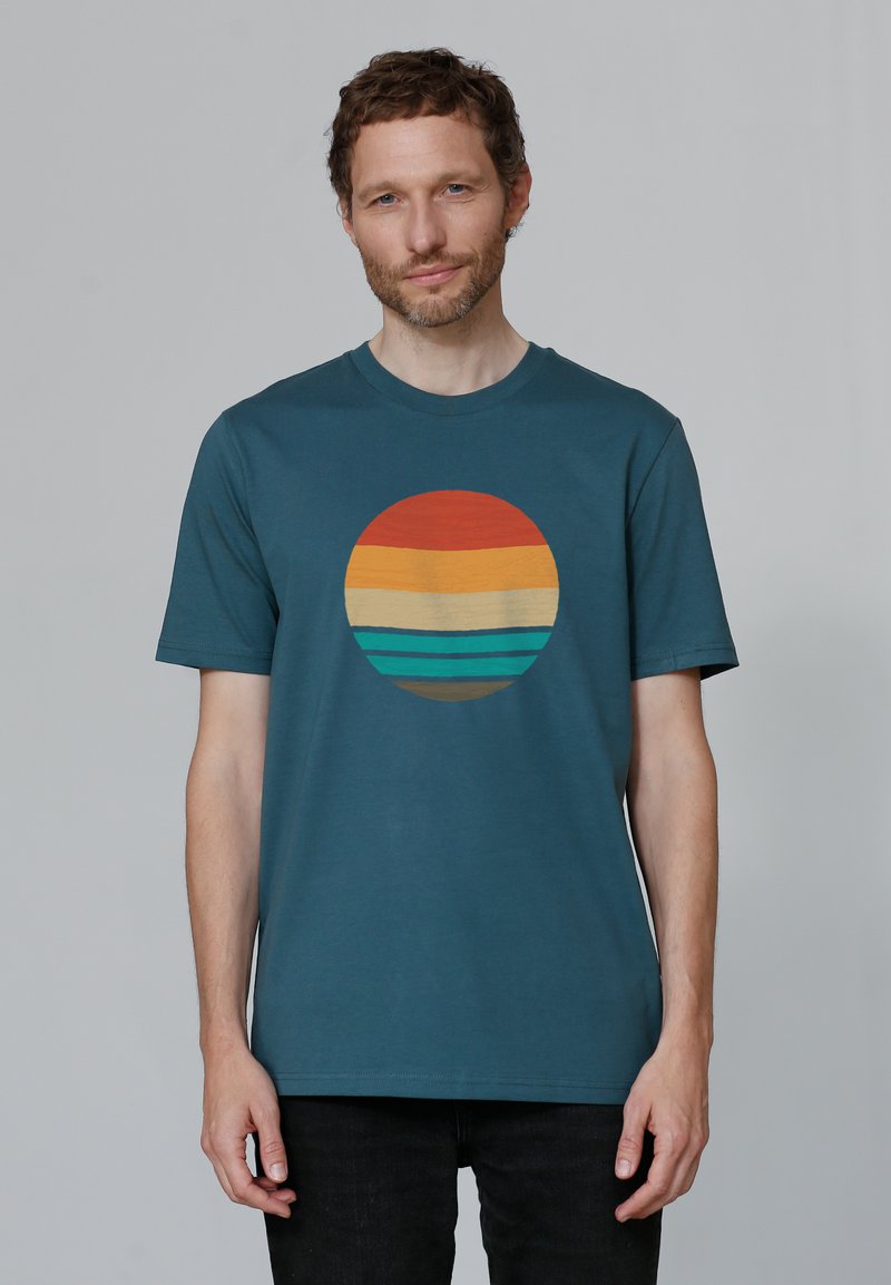 Türaus Baumwoll-T-Shirt in Teal mit einem kreisförmigen Grafikdesign in Orange, Gelb und Teal-Streifen, vor einem schlichten Hintergrund. Kurze Ärmel.
