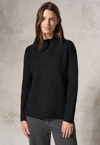 Maglione nero a maglia con un motivo a chevron testurizzato, caratterizzato da un collo alto e polsini e orlo a coste. Indossato sopra pantaloni grigi.