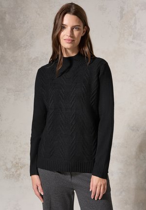 Pull noir en maille avec un motif en chevrons texturé, doté d'un col montant et de poignets et d'un ourlet côtelés. Porté sur un pantalon gris.