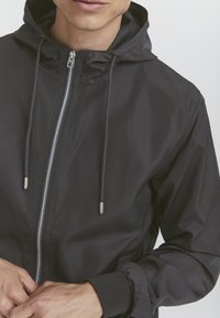 Muž v černé zipové mikině s kapucí a stáhnutelnými šňůrkami, který zapíná zip oběma rukama.