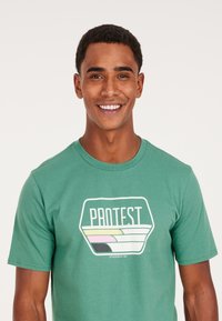 Grøn bomuld t-shirt med et grafisk "PROTEST" logo i hvidt og farverige bølgemønstre. Klassisk rund halsudskæring og korte ærmer.