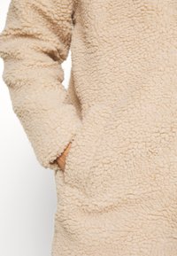 Veste en polaire sherpa beige montrant le tissu texturé et une main glissée dans la poche latérale.