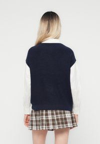 Pull en maille bleu marine à manches courtes, superposé à une chemise blanche à manches longues, associé à une mini-jupe écossaise aux teintes marron et crème.