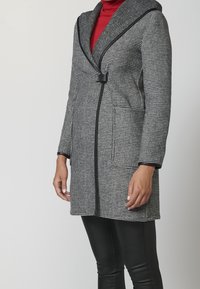 Cappotto grigio e nero a scacchi con cappuccio, dotato di vita aderente e due tasche laterali, realizzato in tessuto testurizzato.