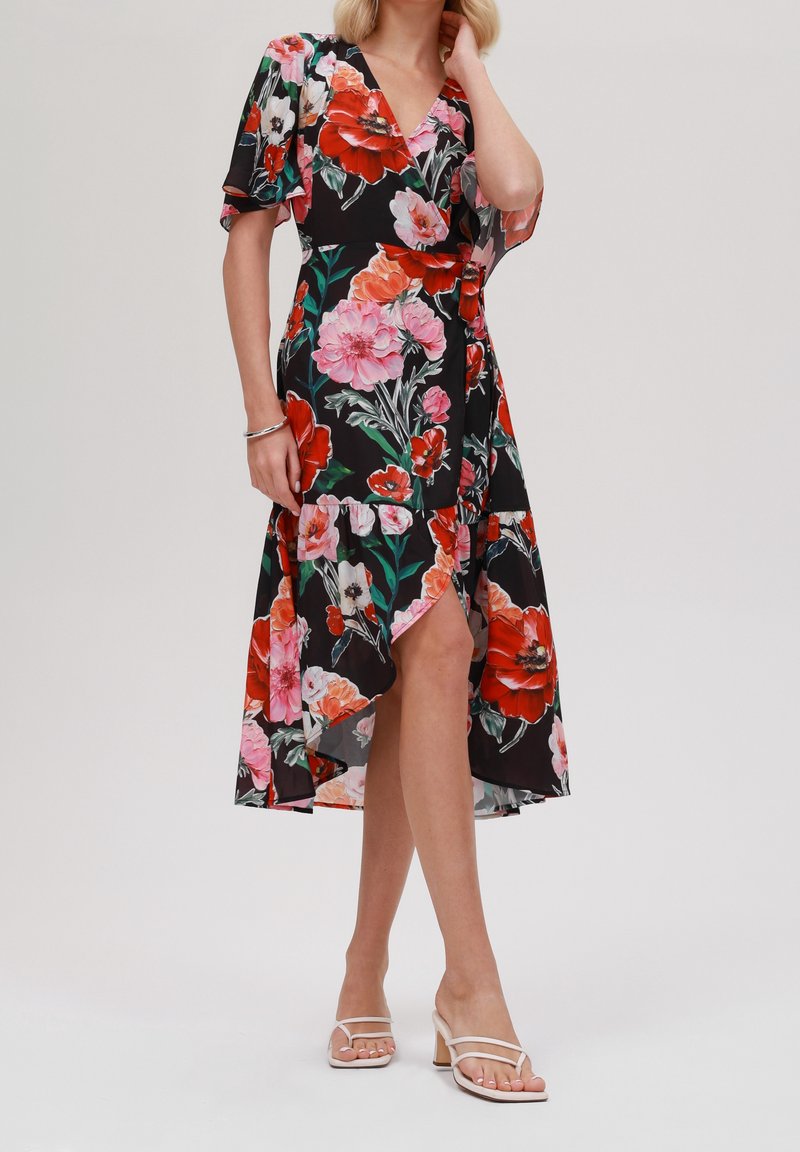 Femme vêtue d'une robe portefeuille noire avec un grand imprimé floral rouge, rose et blanc, des manches volantées, un ourlet asymétrique et des sandales blanches à talons.