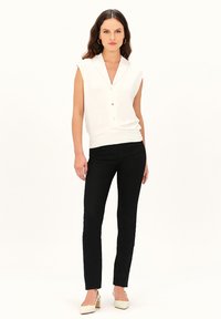 Weiße ärmellose Bluse mit Kragen und Knopfdetails, kombiniert mit schwarzen Slim-Fit-Hosen und weißen High Heels vor einem neutralen Hintergrund.