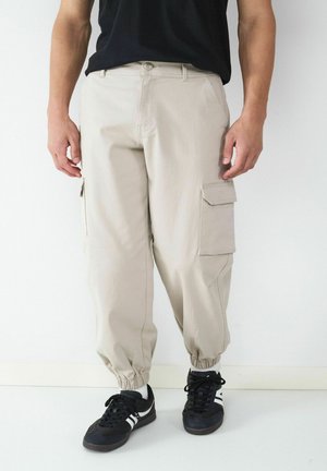 Mężczyzna noszący beżowe spodnie typu cargo z elastycznymi ściągaczami, czarne trampki z białymi paskami oraz czarną koszulkę, stojący przy białej ścianie.